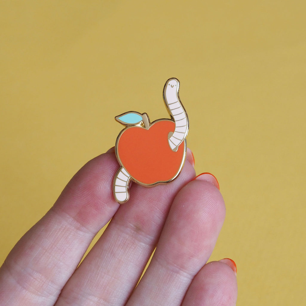 a hand holding an enamel pin of an apple with a worm - nutmeg and arlo